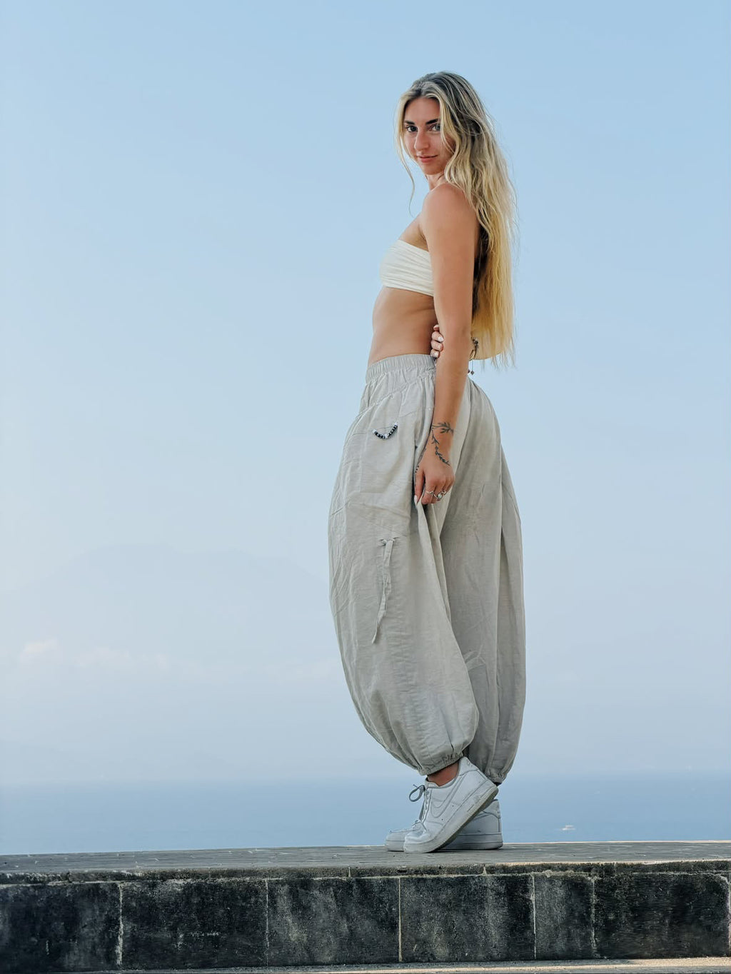 Woman in Linen Love Pants and a white bandeau top on a stone ledge above the ocean, side view