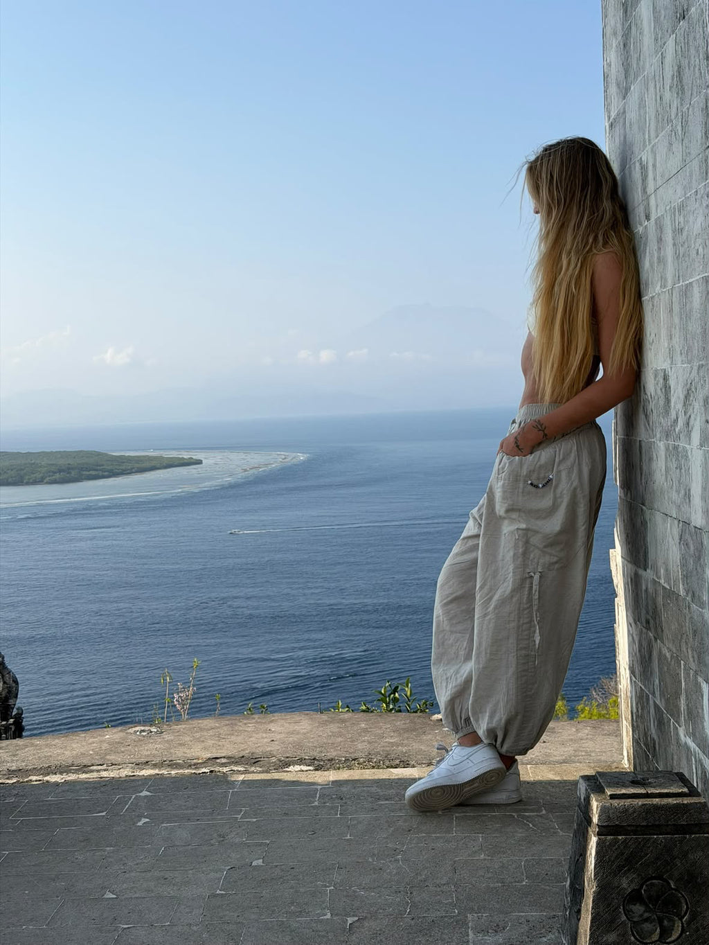 Woman leaning on a stone wall in Linen Love Pants, looking out over the ocean